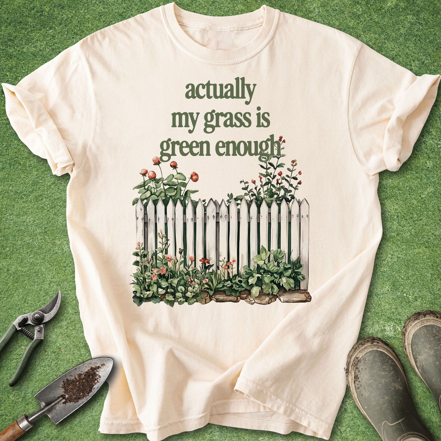A cream-colored short-sleeve t-shirt with a graphic print of a white picket fence and green plants, laid flat on a green grass-like surface with gardening tools and boots nearby.