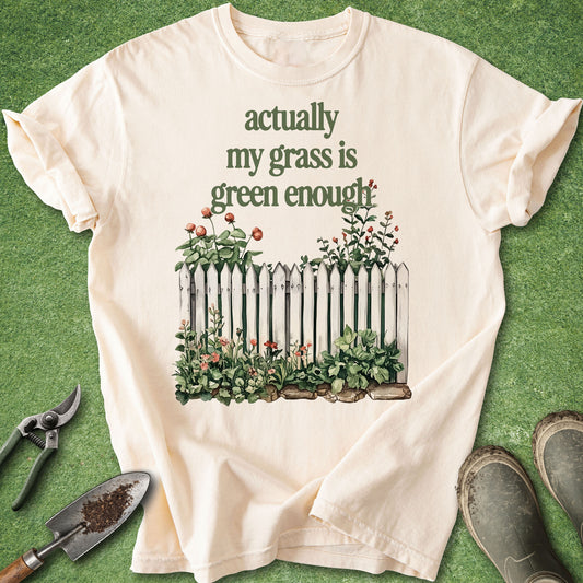 A cream-colored short-sleeve t-shirt with a graphic print of a white picket fence and green plants, laid flat on a green grass-like surface with gardening tools and boots nearby.