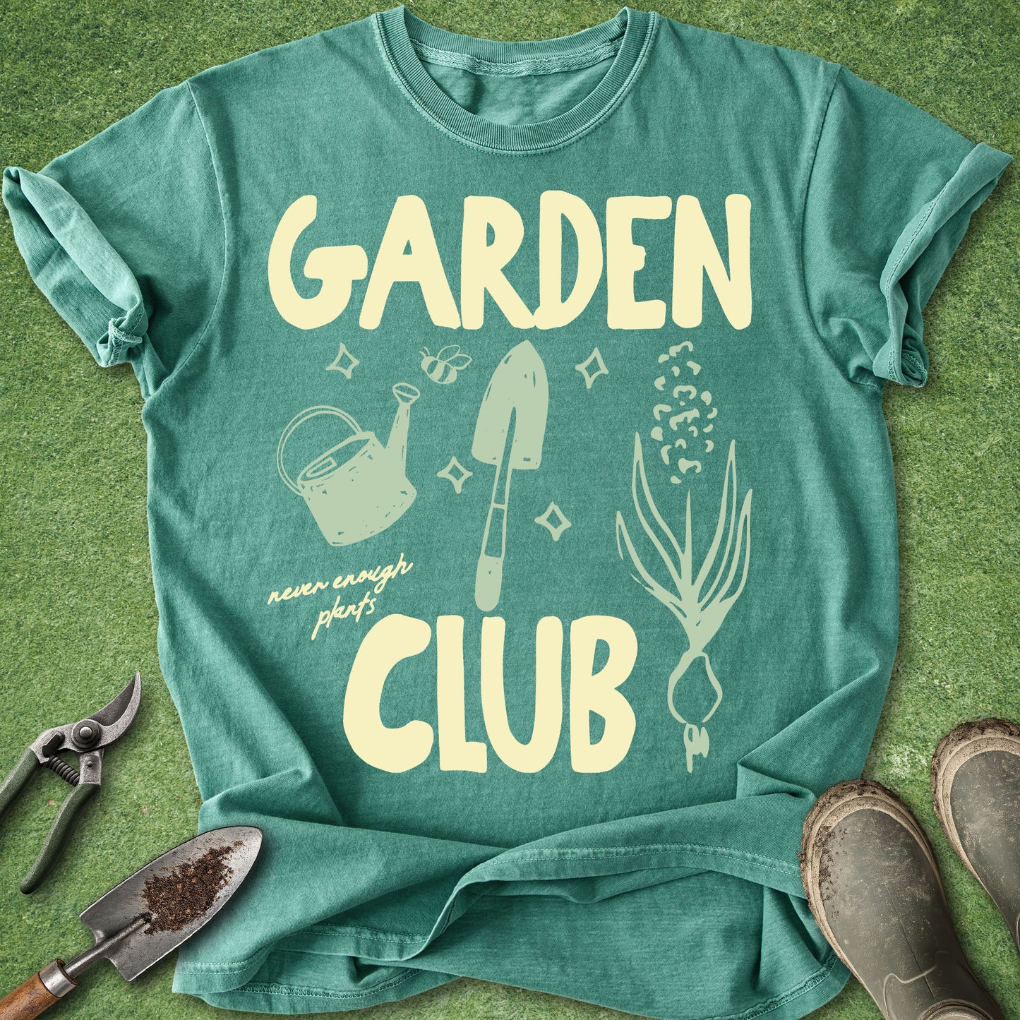 A teal green graphic t-shirt laid flat on a green grass-like surface, surrounded by gardening props including pruning shears, a small trowel, and boots. The shirt features a cream-colored graphic with the words 'GARDEN CLUB' and illustrations of a watering can, trowel, flowers, and a onion.