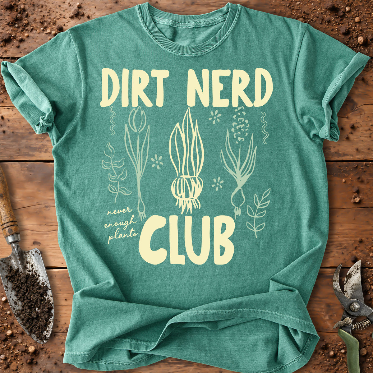 A green graphic t-shirt laid flat on a rustic wooden surface with gardening tools. The t-shirt features a cream-colored graphic that reads 'DIRT NERD CLUB' with illustrations of plants and a onion.