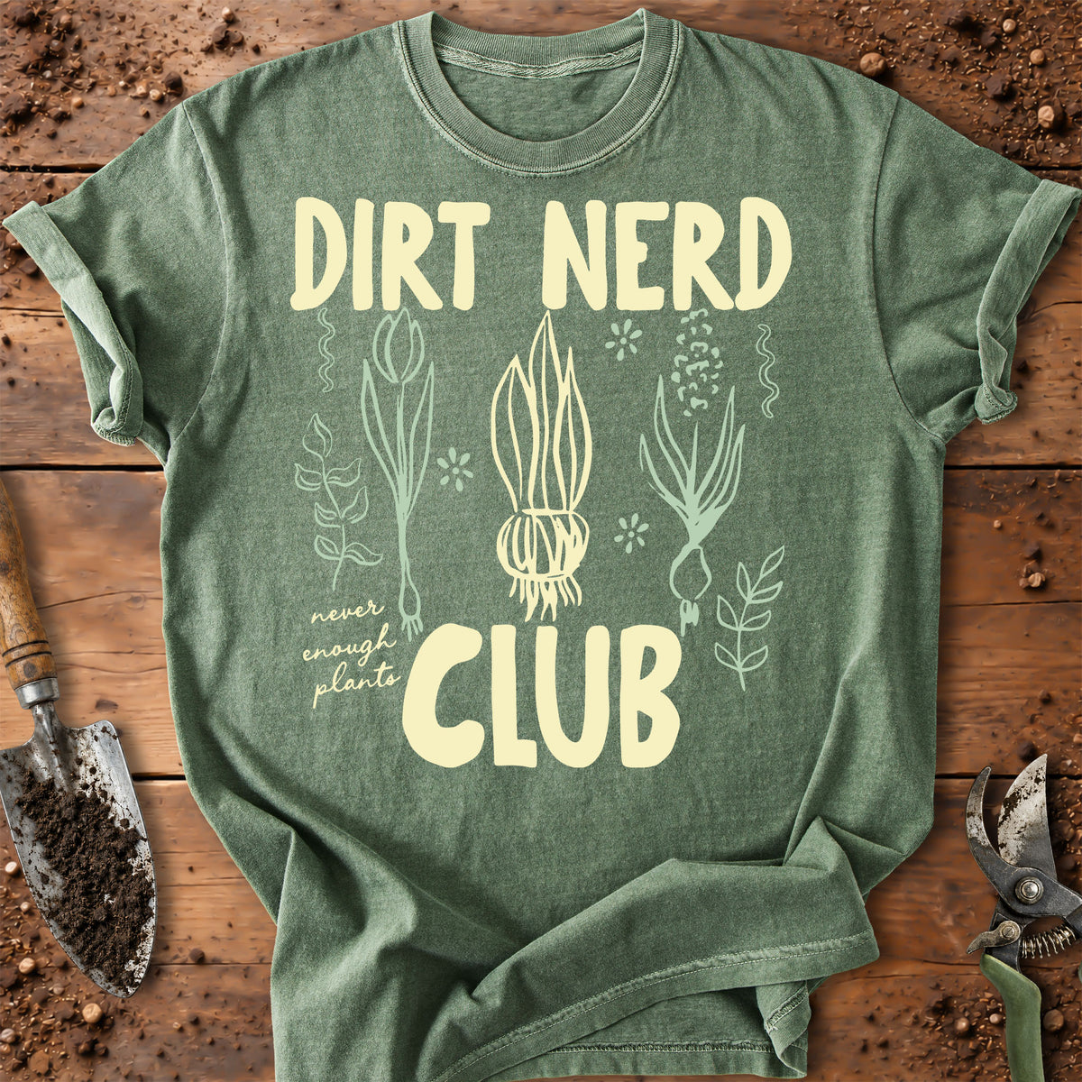 A heather sage green t-shirt with a cream-colored graphic that reads 'DIRT NERD CLUB' and features illustrations of various plants. The shirt is laid flat on a rustic wooden surface with gardening props like a trowel and pruning shears.