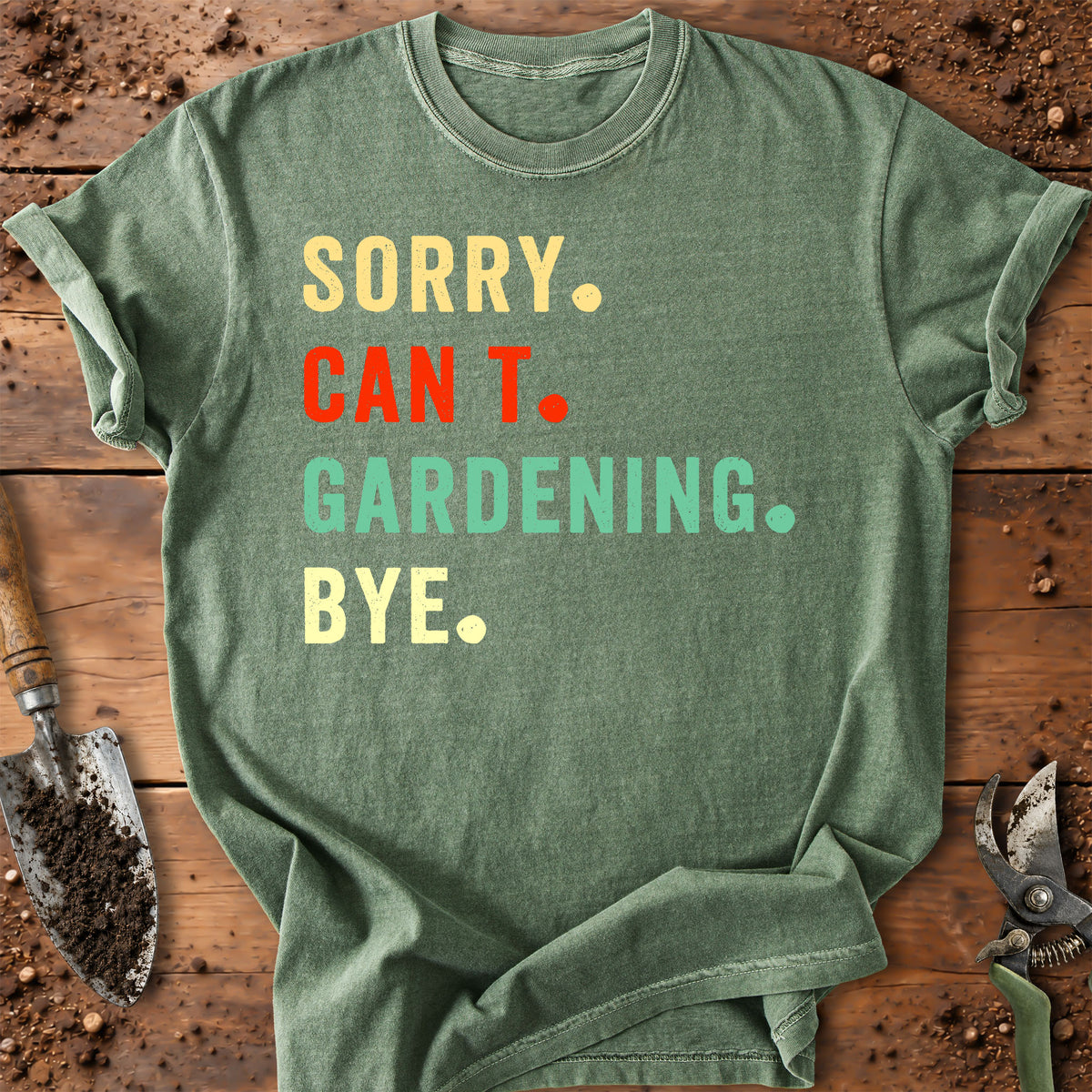 An olive green graphic t-shirt laid flat on a rustic wooden background with gardening props like a trowel and pruning shears. The shirt features the text 'SORRY.