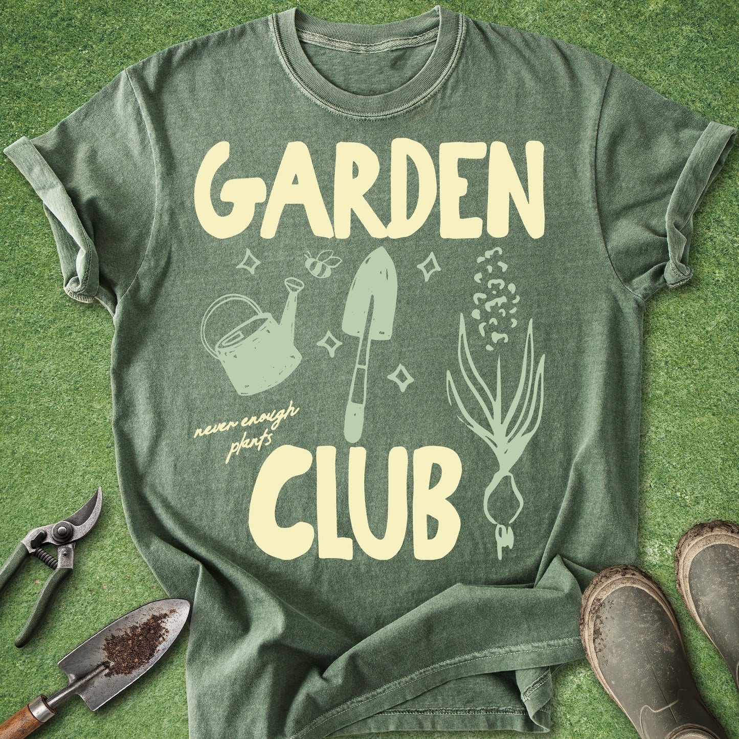 An olive green graphic t-shirt laid flat on a green grass-like surface, surrounded by gardening props including pruning shears, a trowel, and boots. The shirt features a cream-colored graphic that reads 'GARDEN CLUB' with illustrations of a watering can, trowel, flowers, and a plant.