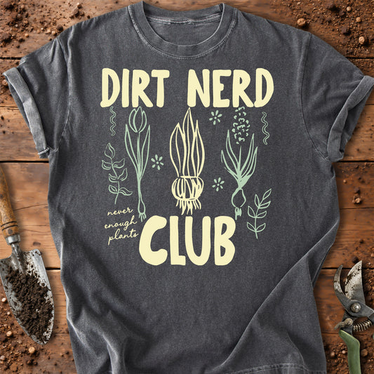 A dark gray graphic t-shirt laid flat on a rustic wooden surface with gardening props like a trowel and pruning shears. The shirt features a cream-colored graphic that reads 'DIRT NERD CLUB' with illustrations of various plants and the phrase 'never enough plants'.
