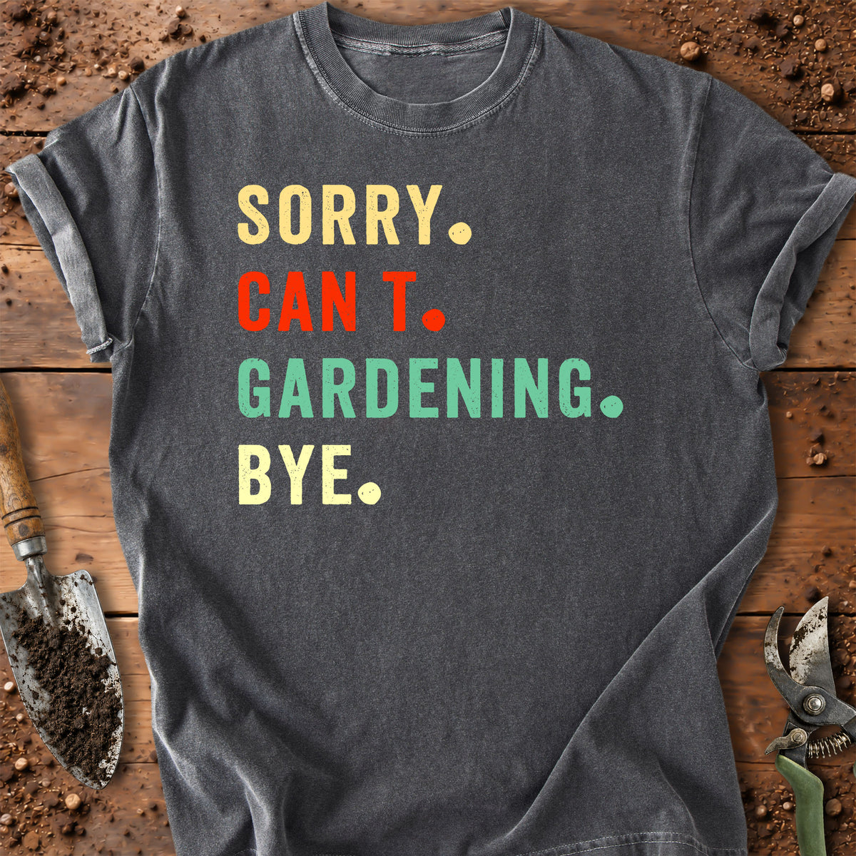 A dark gray graphic t-shirt laid flat on a rustic wooden surface with gardening tools. The shirt features the text 'SORRY.
