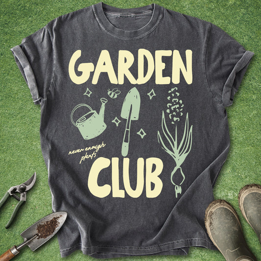 A dark charcoal gray graphic t-shirt laid flat on green grass, featuring the words 'GARDEN CLUB' in a pale yellow font with gardening illustrations including a watering can, trowel, and onion. Gardening tools and boots are styled around the shirt.