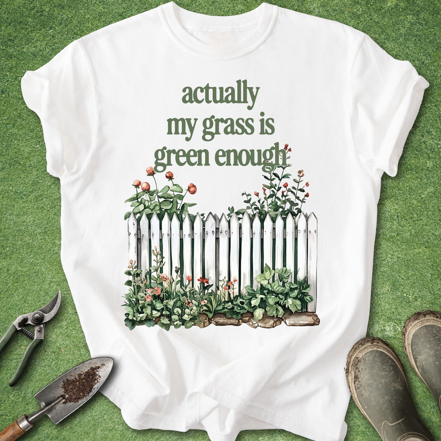 A white graphic t-shirt laid flat on a green grass-like surface, featuring the text 'actually my grass is green enough' above an illustration of a white picket fence and green plants. Gardening tools and muddy boots are placed around the shirt as props.