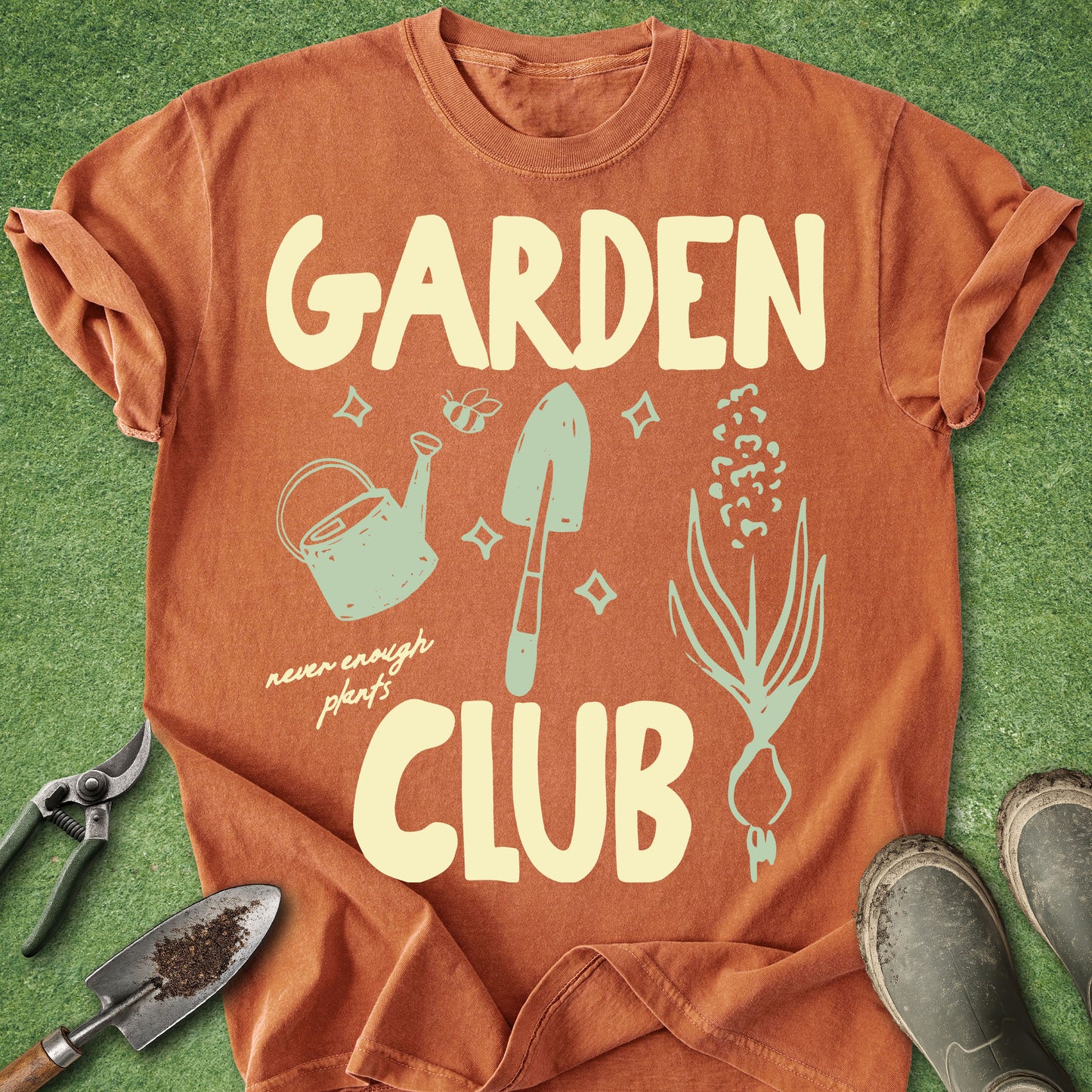 An orange short-sleeve t-shirt laid flat on green grass, featuring a cream-colored graphic that reads 'GARDEN CLUB' with illustrations of a watering can, trowel, seed, and onion. Gardening tools and boots are placed around the shirt as props.