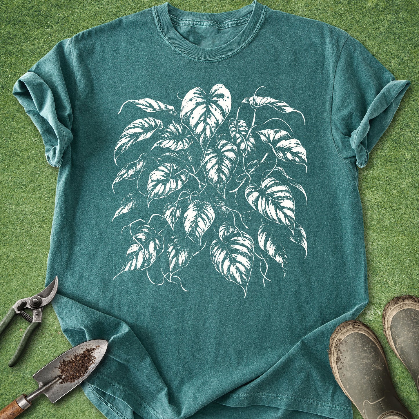 A teal green short-sleeve t-shirt with a white graphic print of a monstera plant, styled in a flat-lay on green grass with gardening tools and boots.