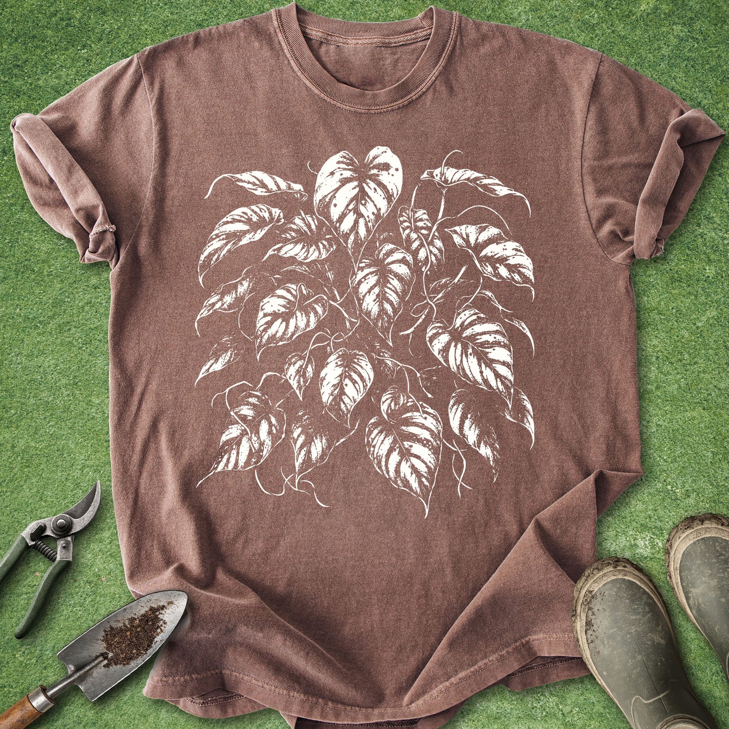 A brown short-sleeve crewneck t-shirt with a white leafy plant graphic, laid flat on green grass with gardening tools and boots nearby.