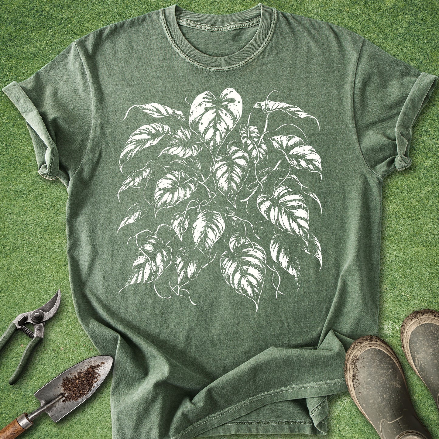 An olive green heathered t-shirt with a white leaf graphic of a houseplant, styled in a flat lay on artificial green grass with gardening tools and boots.