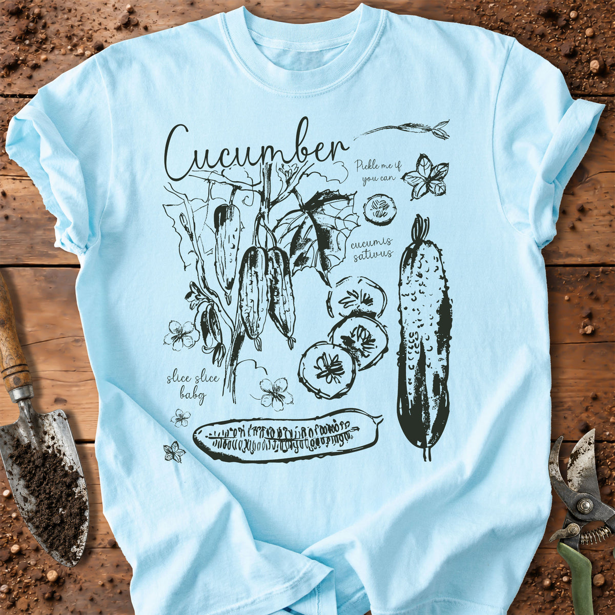 A chambray graphic t-shirt featuring black ink illustrations of cucumbers, leaves, and text, laid flat on a rustic wooden surface with gardening tools.