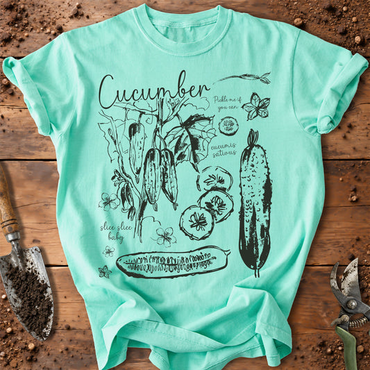A mint green graphic t-shirt featuring black ink illustrations of cucumbers, leaves, and text, laid flat on a rustic wooden surface with gardening tools.