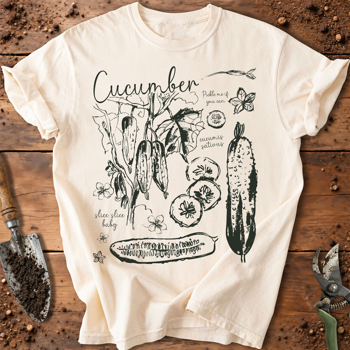 An off-white short-sleeve t-shirt with a black hand-drawn style graphic of cucumbers, leaves, and text, laid flat on a rustic wooden surface with gardening tools.