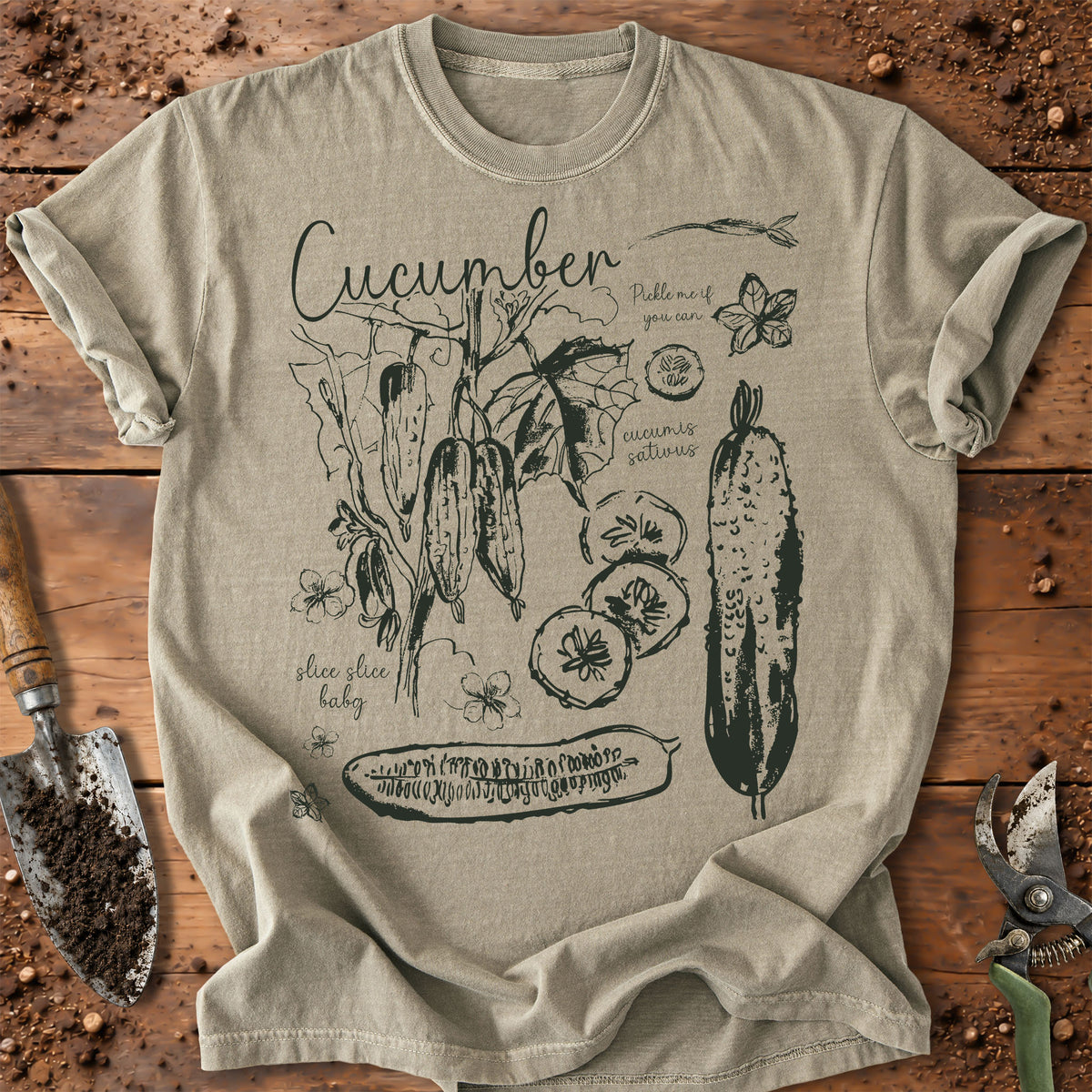 A light beige graphic t-shirt featuring a dark green cucumber-themed illustration, laid flat on a rustic wooden surface with gardening tools.