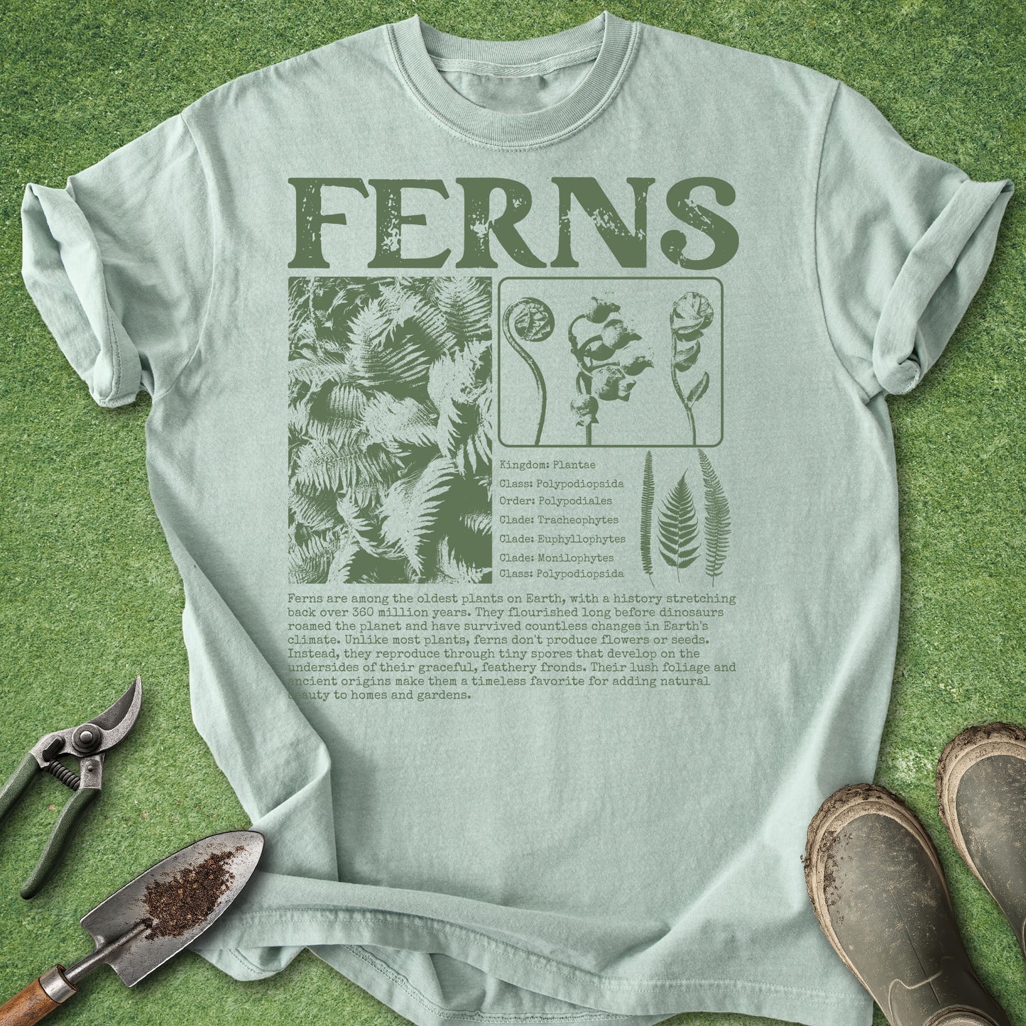 A light sage green t-shirt with a dark green 'FERNS' graphic print, laid flat on a green grass-like background with gardening props like pruning shears, a trowel, and boots.