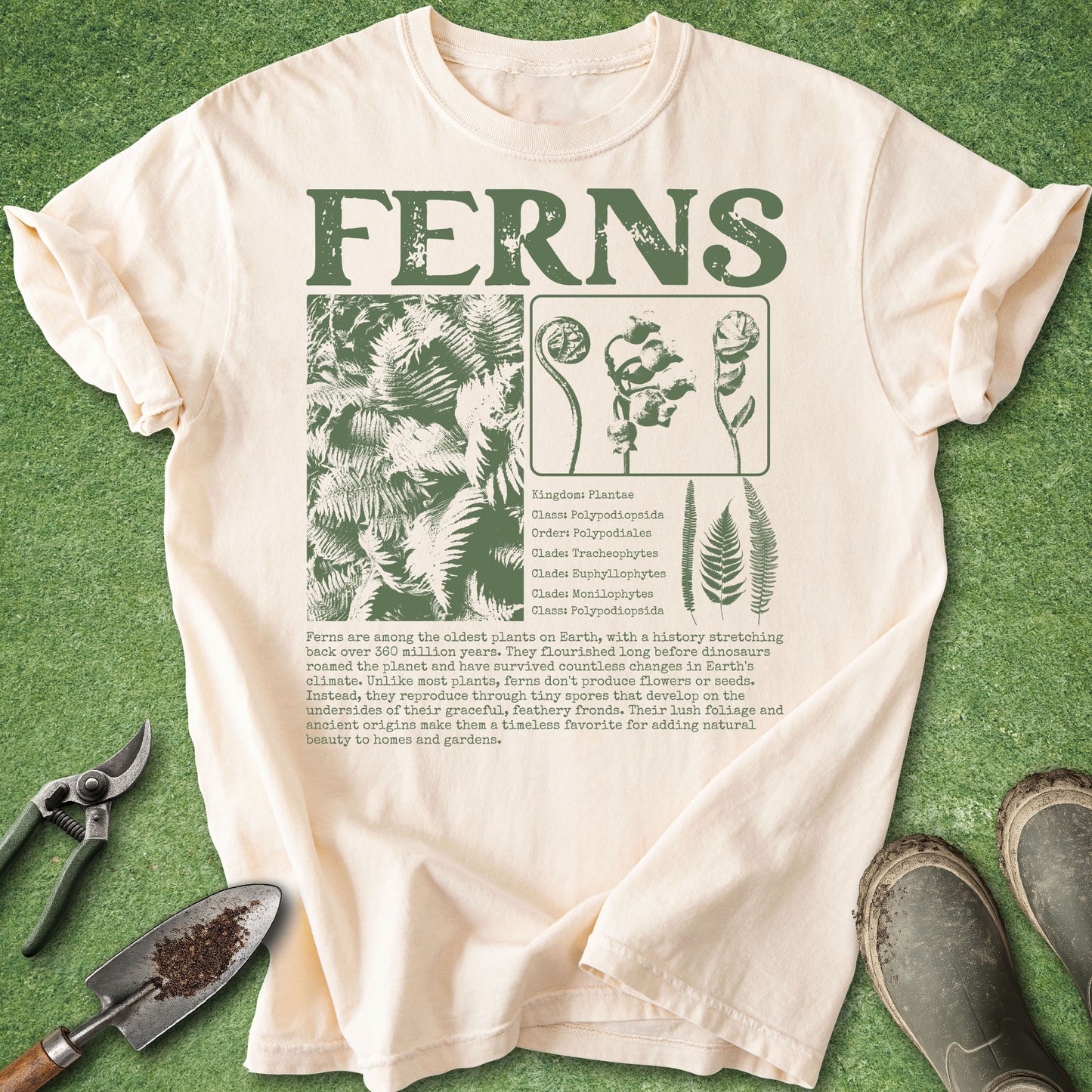 A cream-colored short-sleeve t-shirt with a green graphic print reading 'FERNS' and featuring botanical illustrations and gardening tools, laid flat on a green grass-like background with gardening shears, a trowel, and boots nearby.