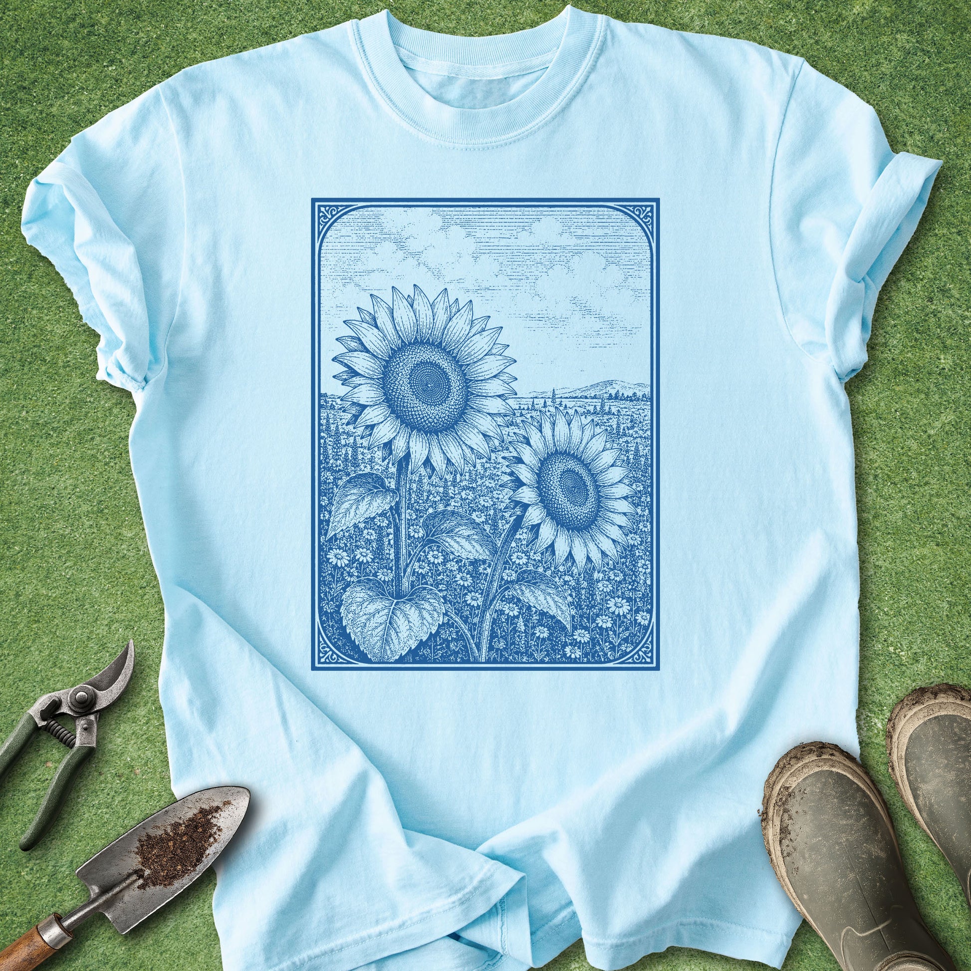 A light blue short-sleeve crewneck t-shirt laid flat on green grass, featuring a blue line-art graphic of two sunflowers. Gardening tools and boots are styled around the shirt.
