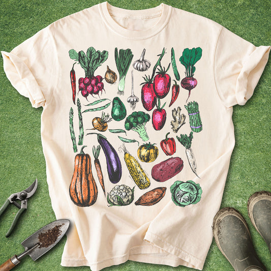 Ivory Comfort Colors t-shirt featuring a colorful collection of hand-drawn vegetables including tomatoes, peppers, radishes, squash, cabbage, broccoli, carrots, potatoes, garlic, asparagus, and eggplant in a vintage botanical style.