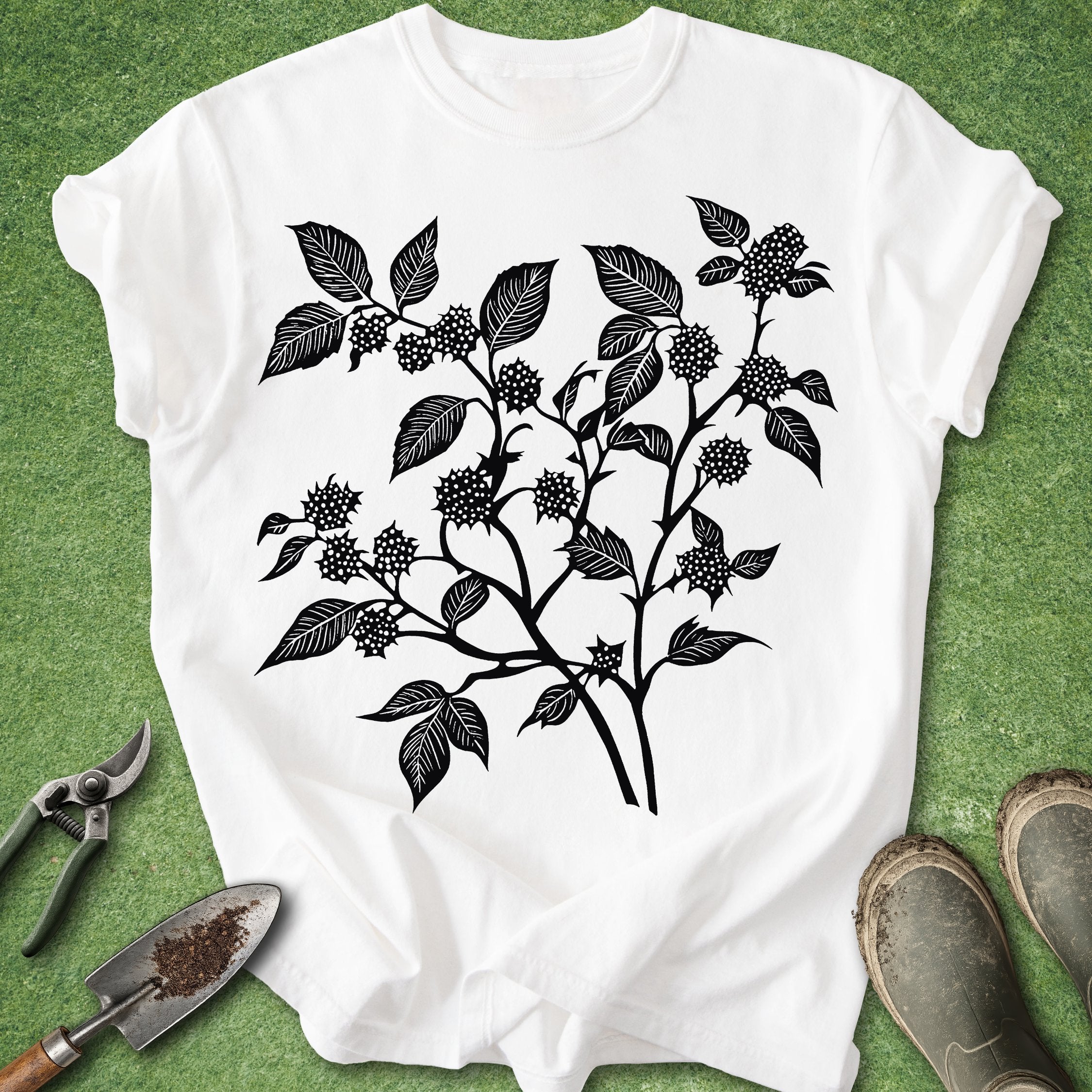 White t-shirt with black leaf and berry design on a green background with gardening tools.
