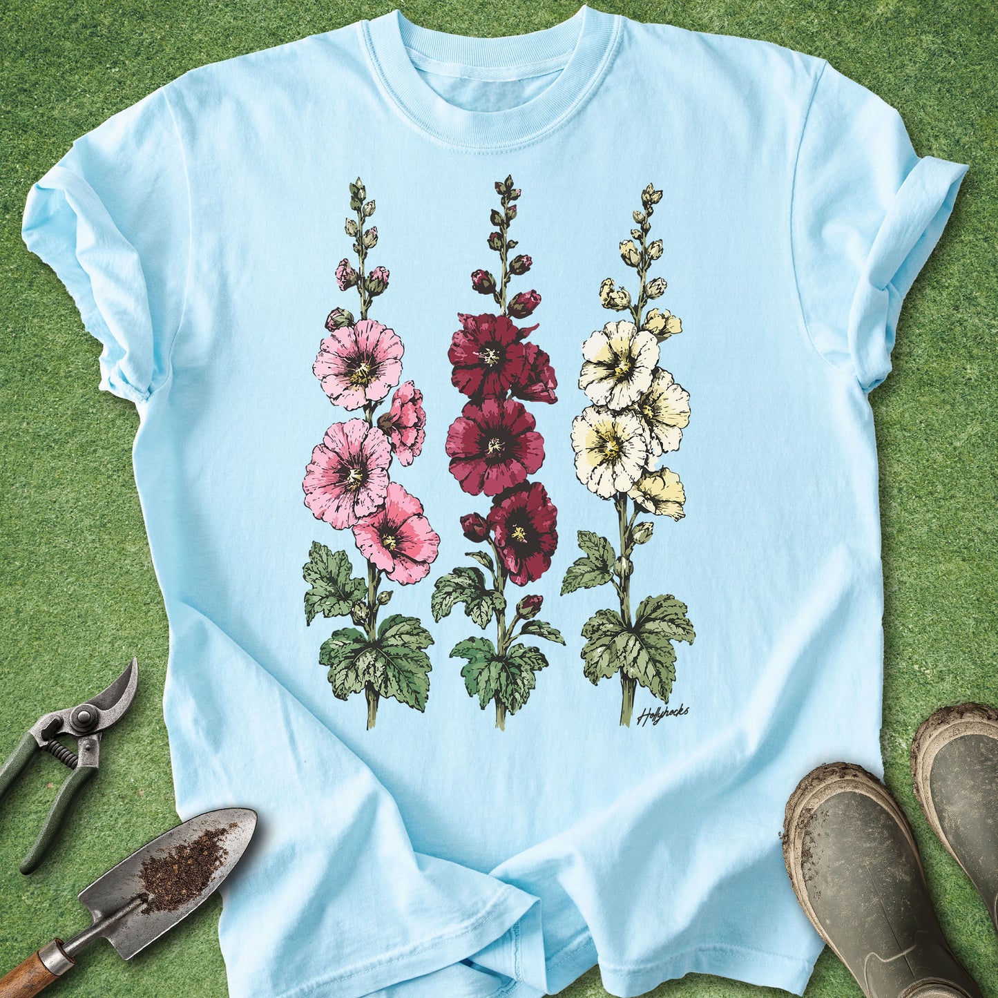 Chambray Comfort Colors t-shirt featuring detailed botanical illustrations of three blooming hollyhock flowers in pink, burgundy, and white with green foliage in a vintage botanical style.