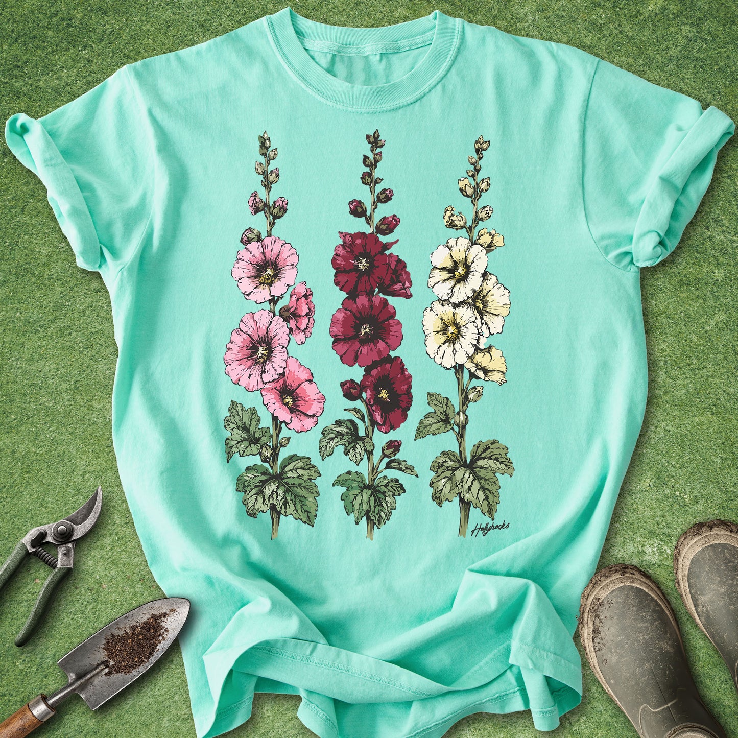 Island Reef Comfort Colors t-shirt featuring detailed botanical illustrations of three blooming hollyhock flowers in pink, burgundy, and white with green foliage in a vintage botanical style.