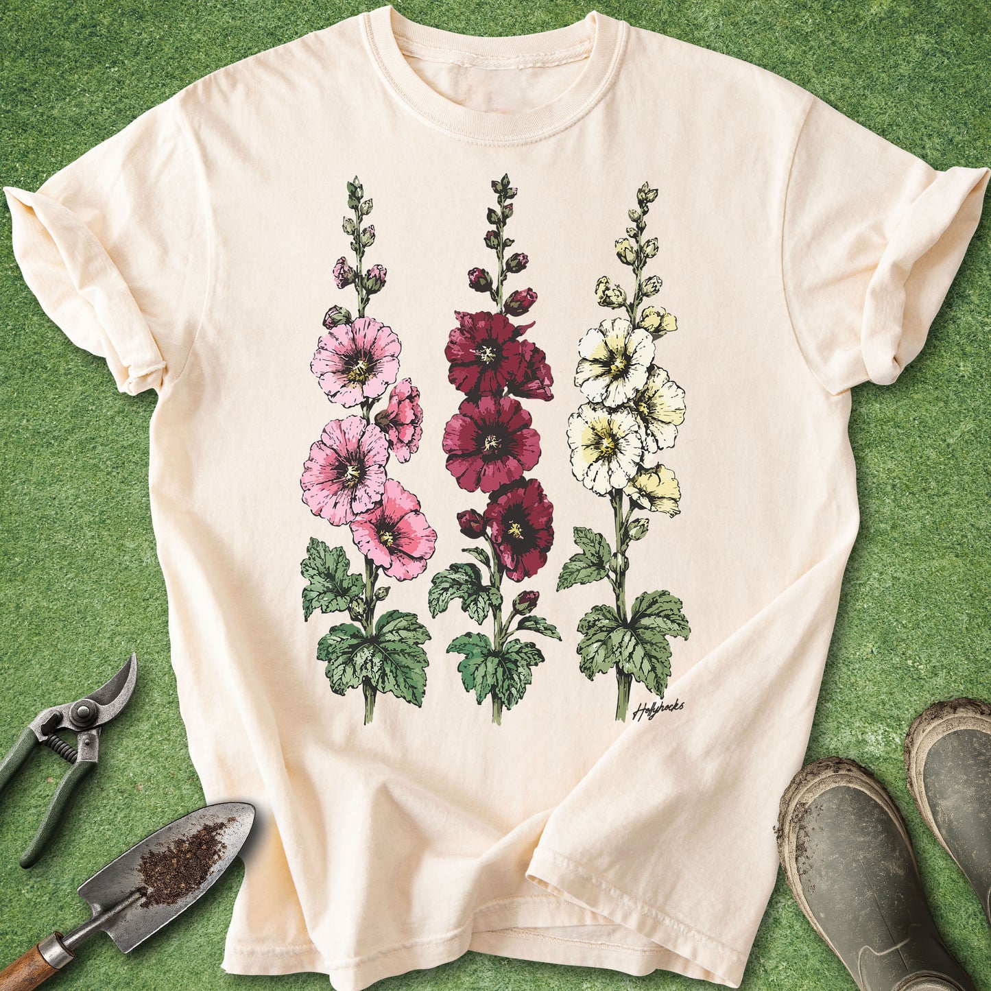 Ivory Comfort Colors t-shirt featuring detailed botanical illustrations of three blooming hollyhock flowers in pink, burgundy, and white with green foliage in a vintage botanical style.