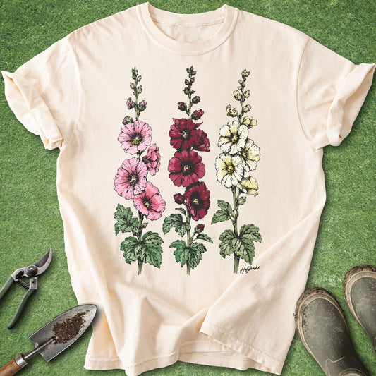 Ivory Comfort Colors t-shirt featuring detailed botanical illustrations of three blooming hollyhock flowers in pink, burgundy, and white with green foliage in a vintage botanical style.