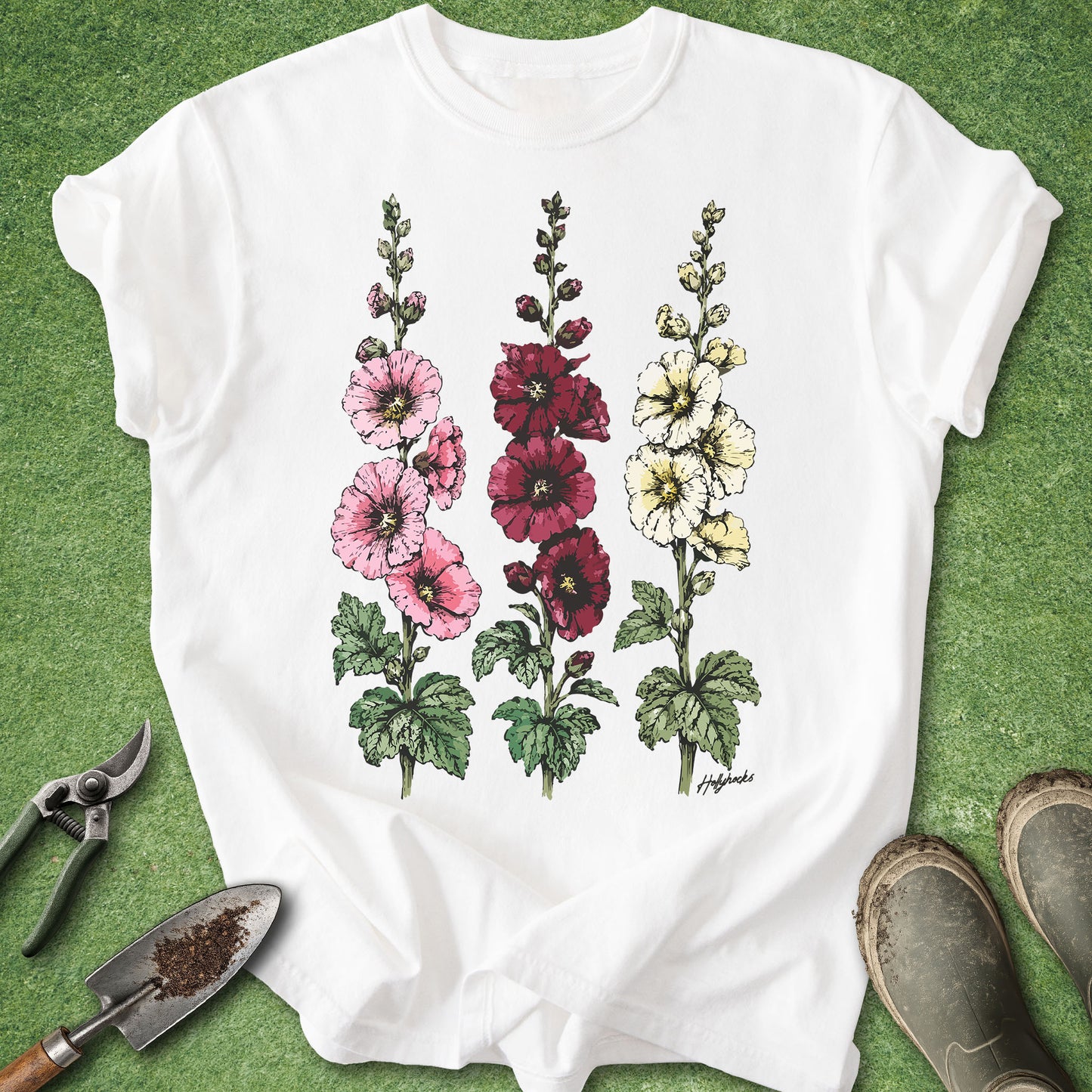 White Comfort Colors t-shirt featuring detailed botanical illustrations of three blooming hollyhock flowers in pink, burgundy, and white with green foliage in a vintage botanical style.