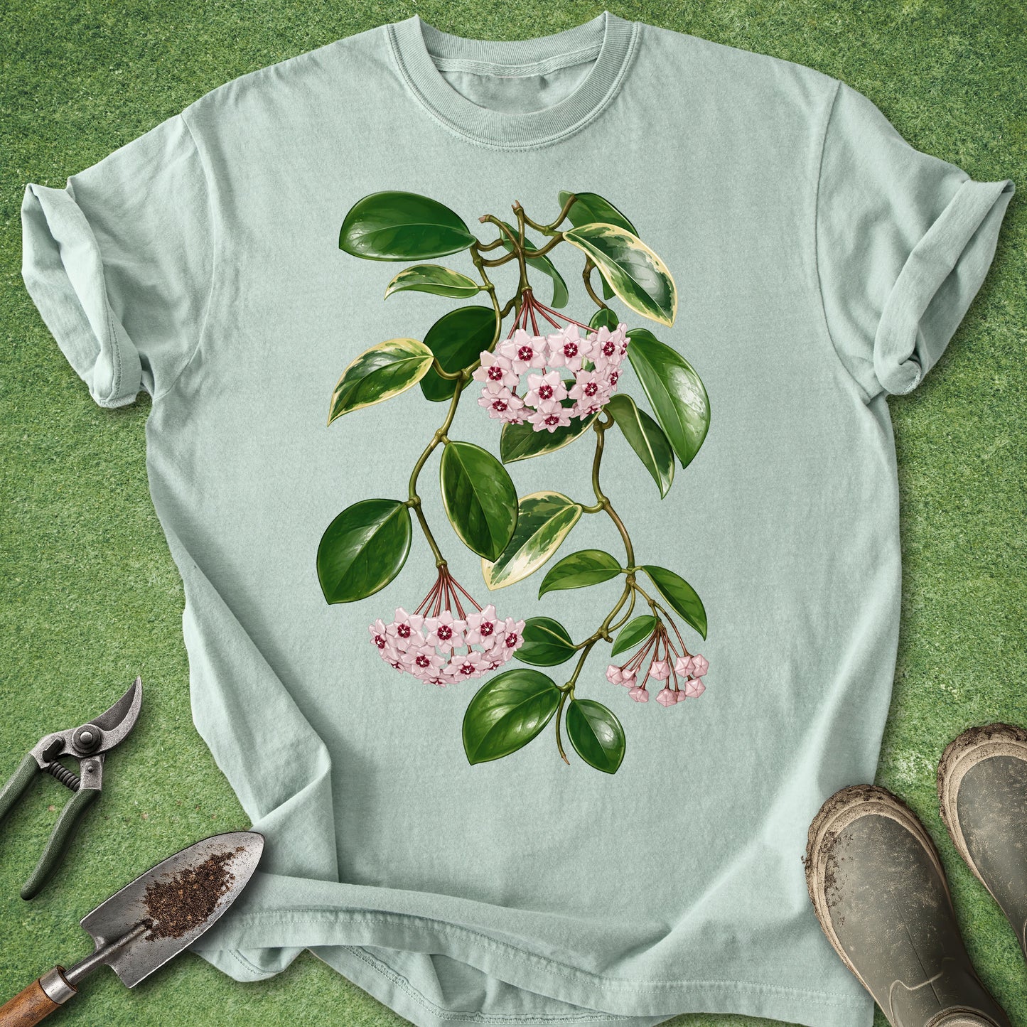 Green t-shirt featuring a botanical Hoya Carnosa wax plant with trailing vines, glossy green and variegated leaves, and clusters of pink star-shaped flowers.