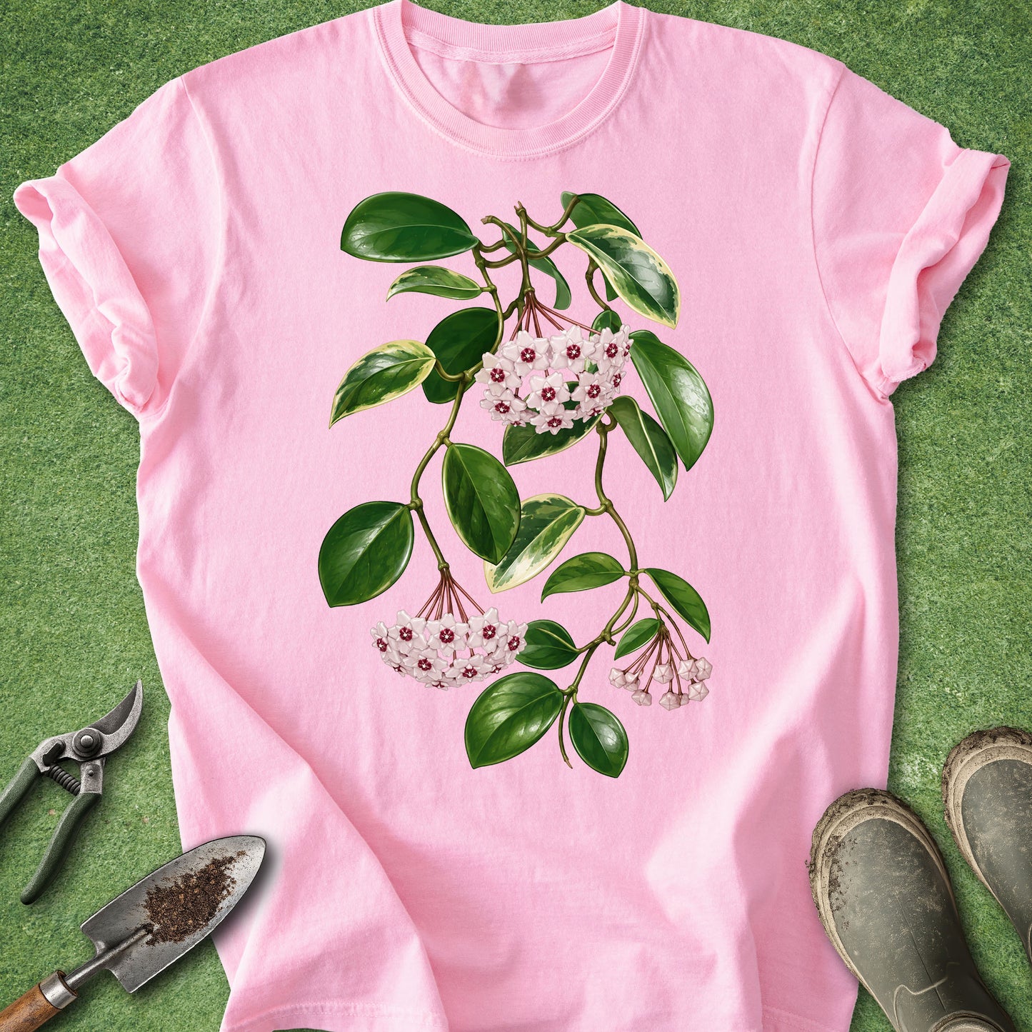 A pink short-sleeve crewneck t-shirt featuring a botanical illustration of a green leafy branch with small white and pink flowers. The shirt is laid flat on a green grass-like surface, styled with gardening tools (shears, trowel) and muddy boots in the corners.