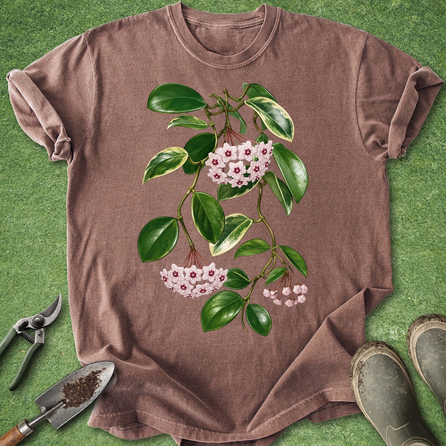 A brown short-sleeve crewneck t-shirt featuring a botanical graphic of a hollyhock branch with green leaves and pink flowers, laid flat on a green grass-like background with gardening tools and boots nearby.