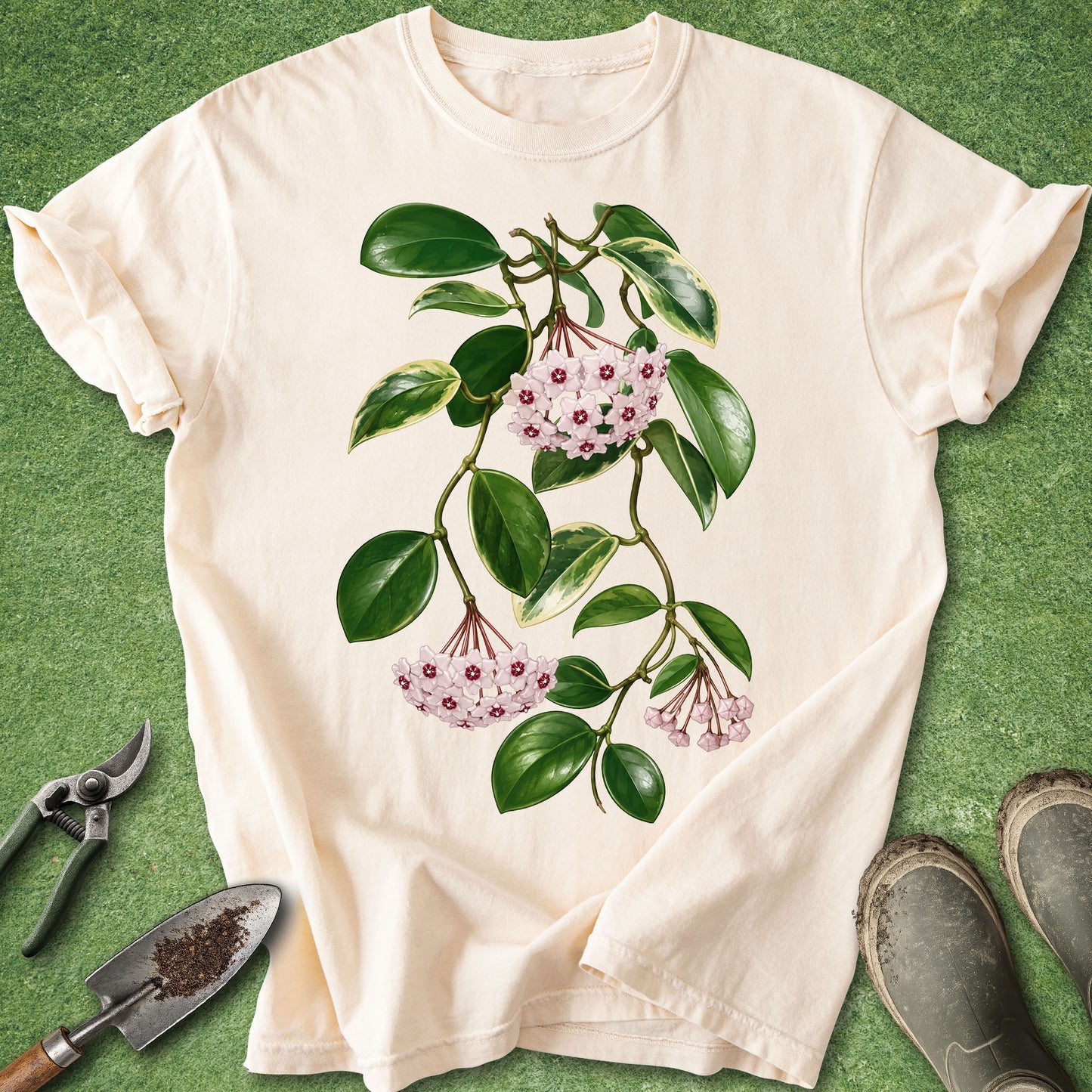 A cream-colored short-sleeve t-shirt with a botanical graphic of green leaves and pink flowers, laid flat on a green grass-like background with gardening tools and boots nearby.