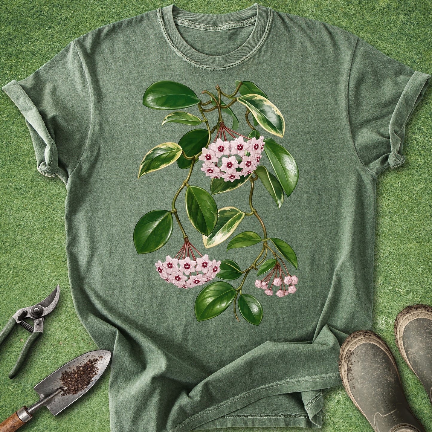An olive green short-sleeve t-shirt with a botanical graphic of a flowering branch, laid flat on a green grass-like surface with gardening tools and boots nearby.