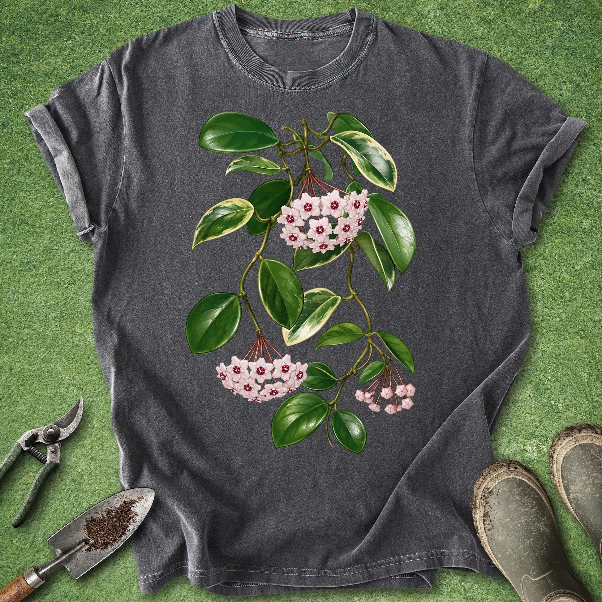 A dark gray t-shirt with a botanical print of green leaves and pink flowers, styled flat on a green grass-like background with gardening tools and boots.