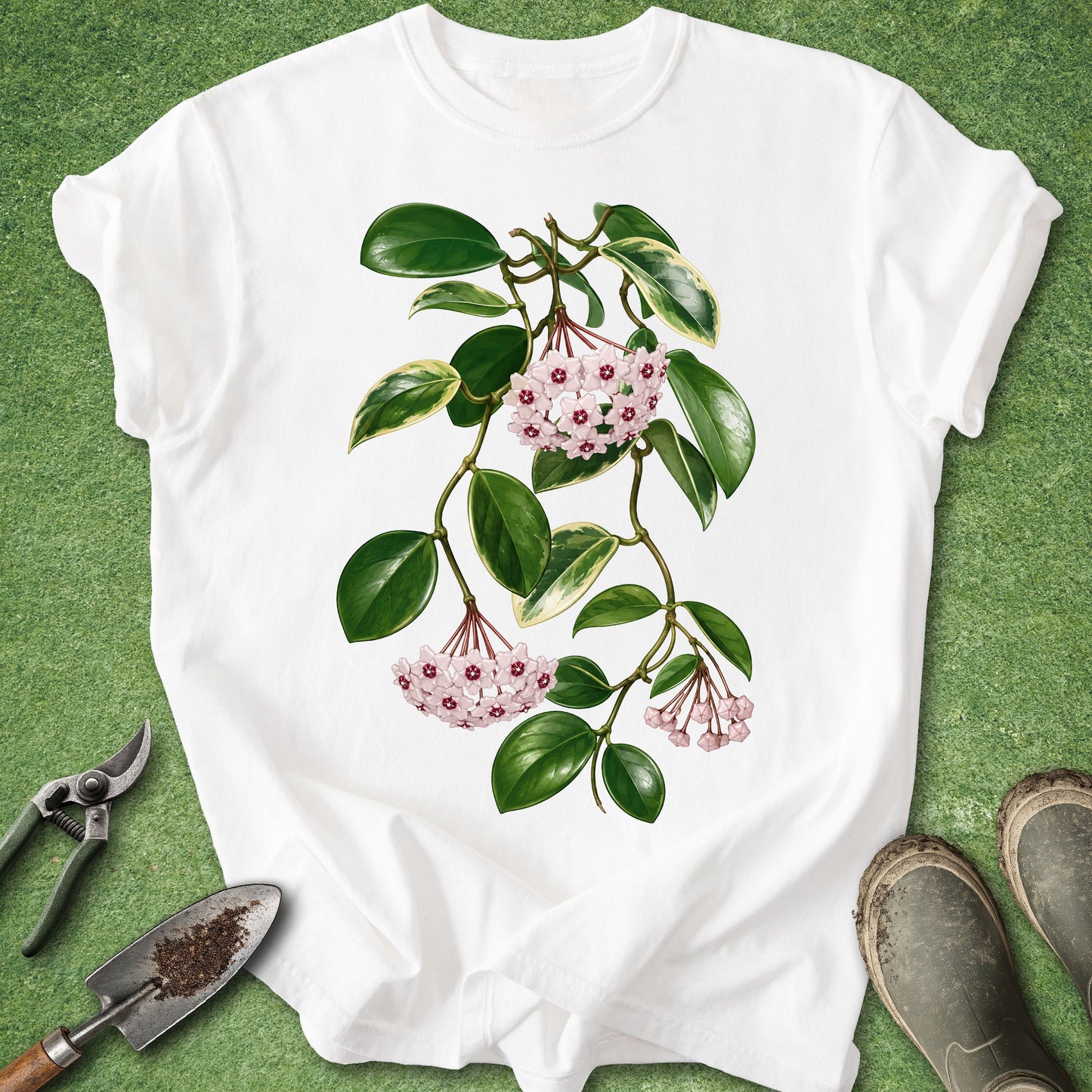 A white short-sleeve T-shirt laid flat on a green grass-like surface, featuring a botanical print of green leaves and pink flowers. Gardening tools and muddy boots are placed around the shirt as styled props.