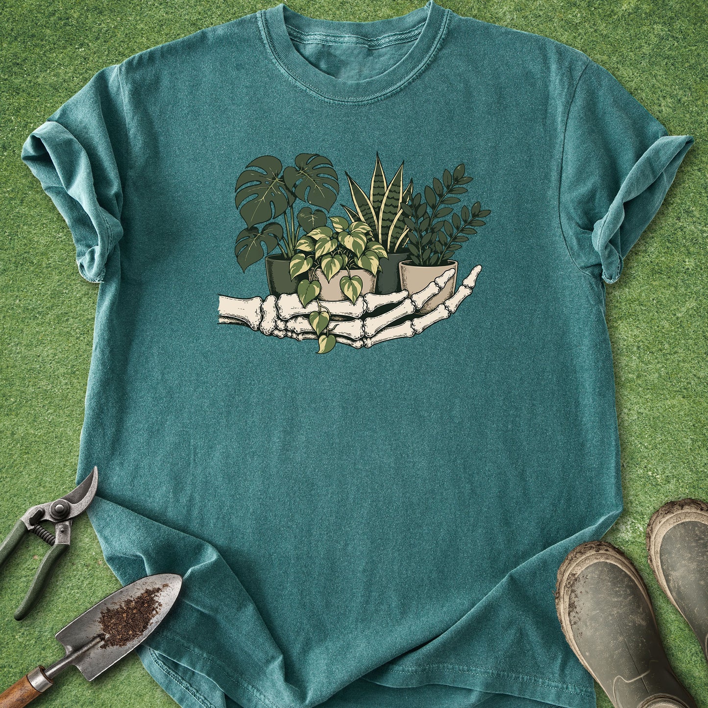 A teal short-sleeve t-shirt laid flat on green artificial grass, featuring a graphic of skeletal hands holding potted houseplants. Gardening props, including pruning shears, a trowel, and boots, are arranged around the shirt.