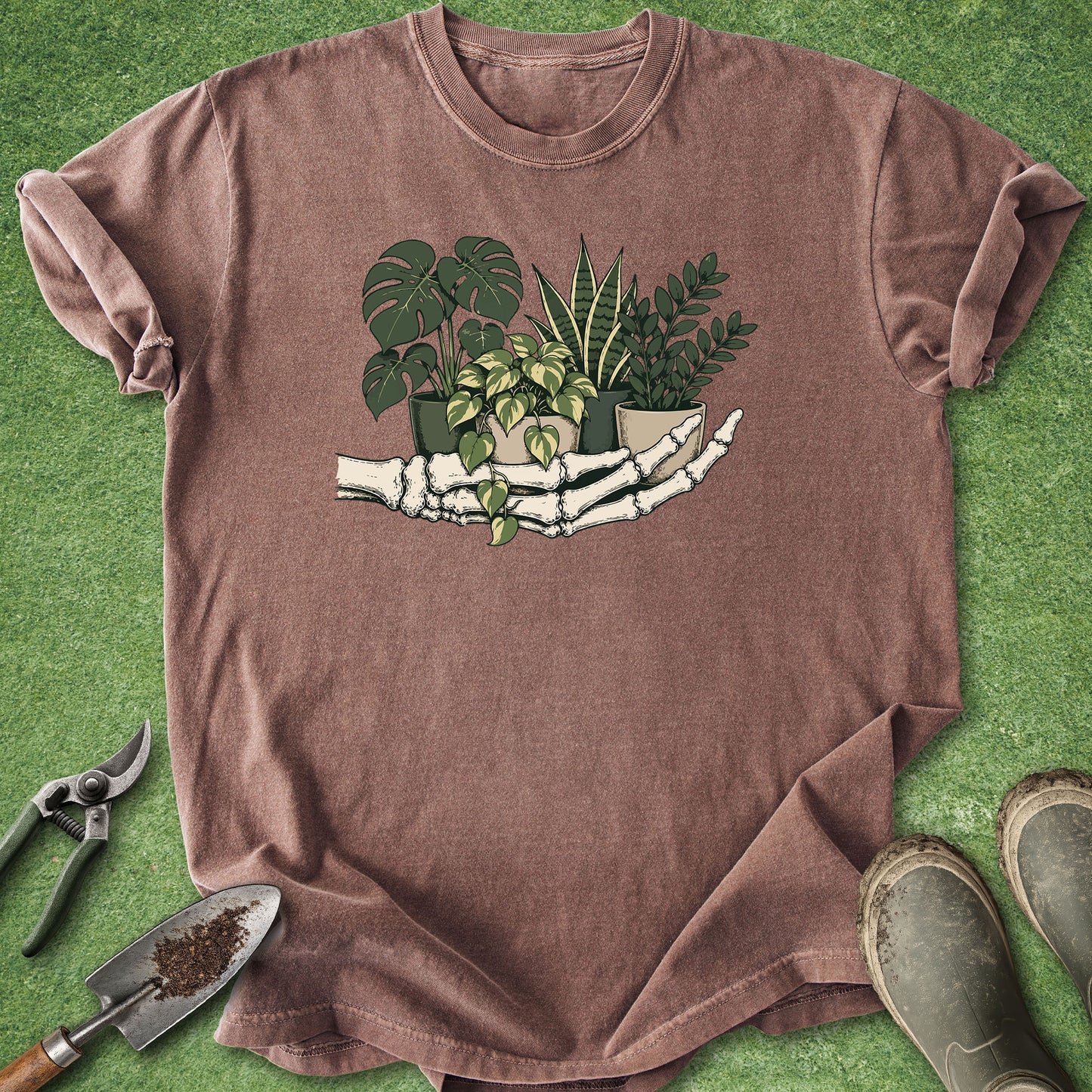 A brown short-sleeve t-shirt laid flat on green artificial grass, featuring a graphic of a skeleton hand holding several potted green plants. Gardening props, including pruning shears, a trowel with soil, and gardening boots, are arranged around the shirt.