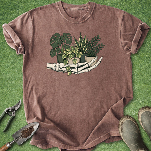 A brown short-sleeve t-shirt laid flat on green artificial grass, featuring a graphic of a skeleton hand holding several potted green plants. Gardening props, including pruning shears, a trowel with soil, and gardening boots, are arranged around the shirt.