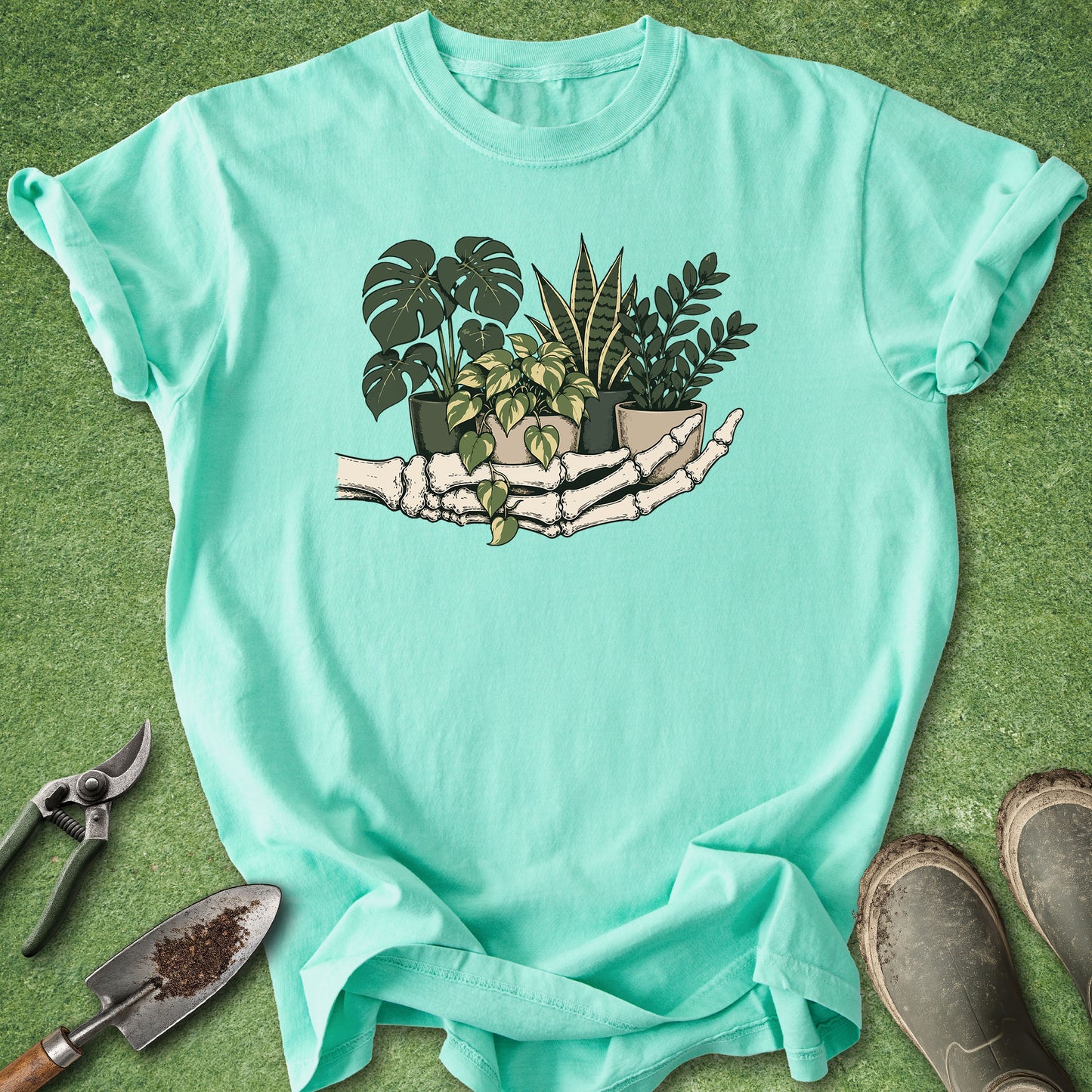 A mint green short-sleeve t-shirt laid flat on a green grass-like surface, featuring a graphic of skeleton hands holding potted plants. Gardening props including pruning shears, a trowel, and boots are arranged around the shirt.