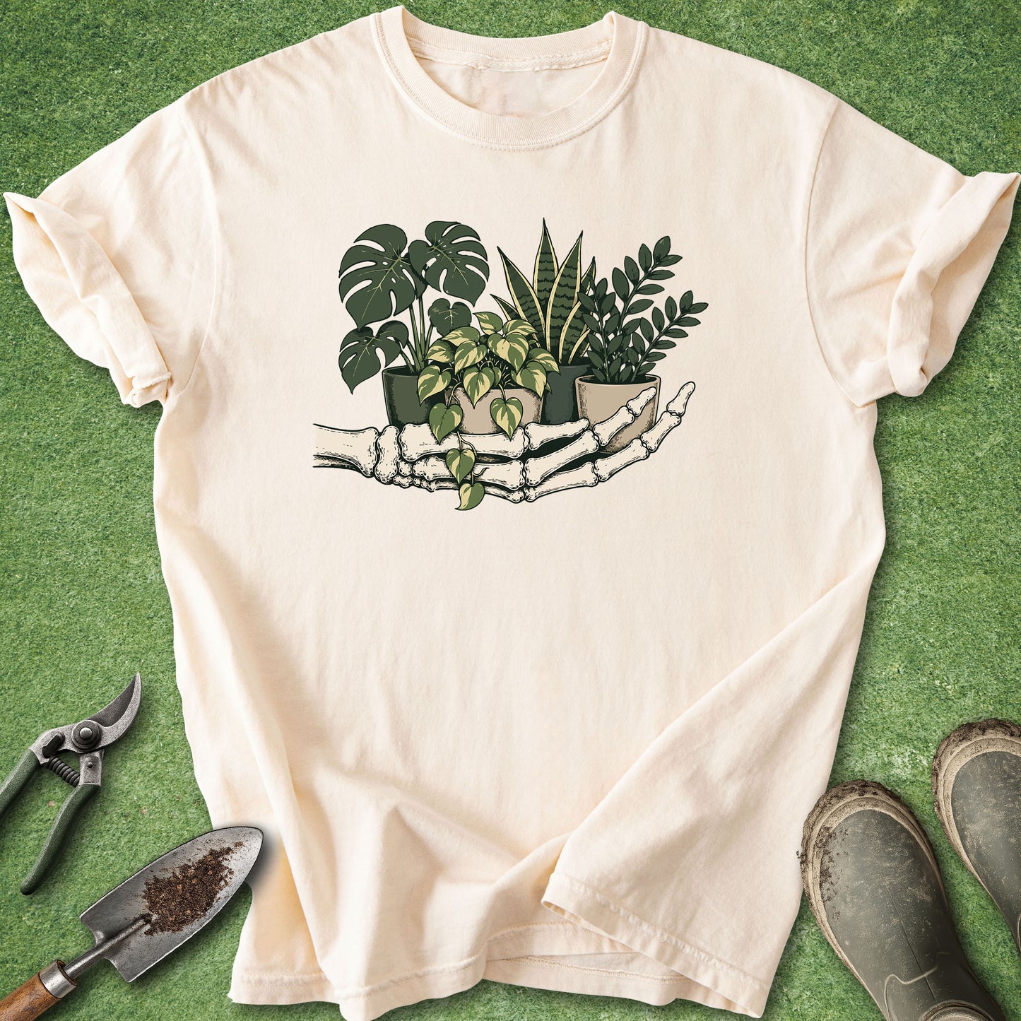 A cream-colored short-sleeve t-shirt with a graphic of a skeleton hand holding potted houseplants, laid flat on a green grass-like surface with gardening tools and shoes nearby.
