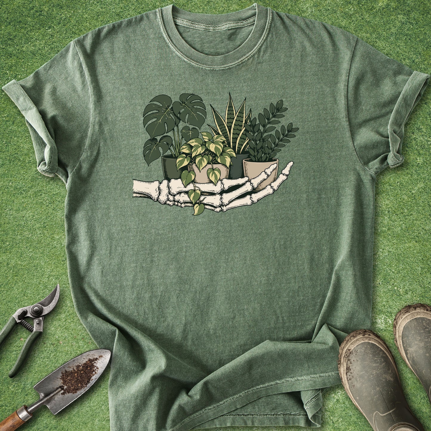 An olive green short-sleeve t-shirt laid flat on a green grass-like surface, featuring a graphic of skeletal hands holding several potted green plants. Gardening props, including pruning shears, a trowel, and boots, are arranged around the shirt.