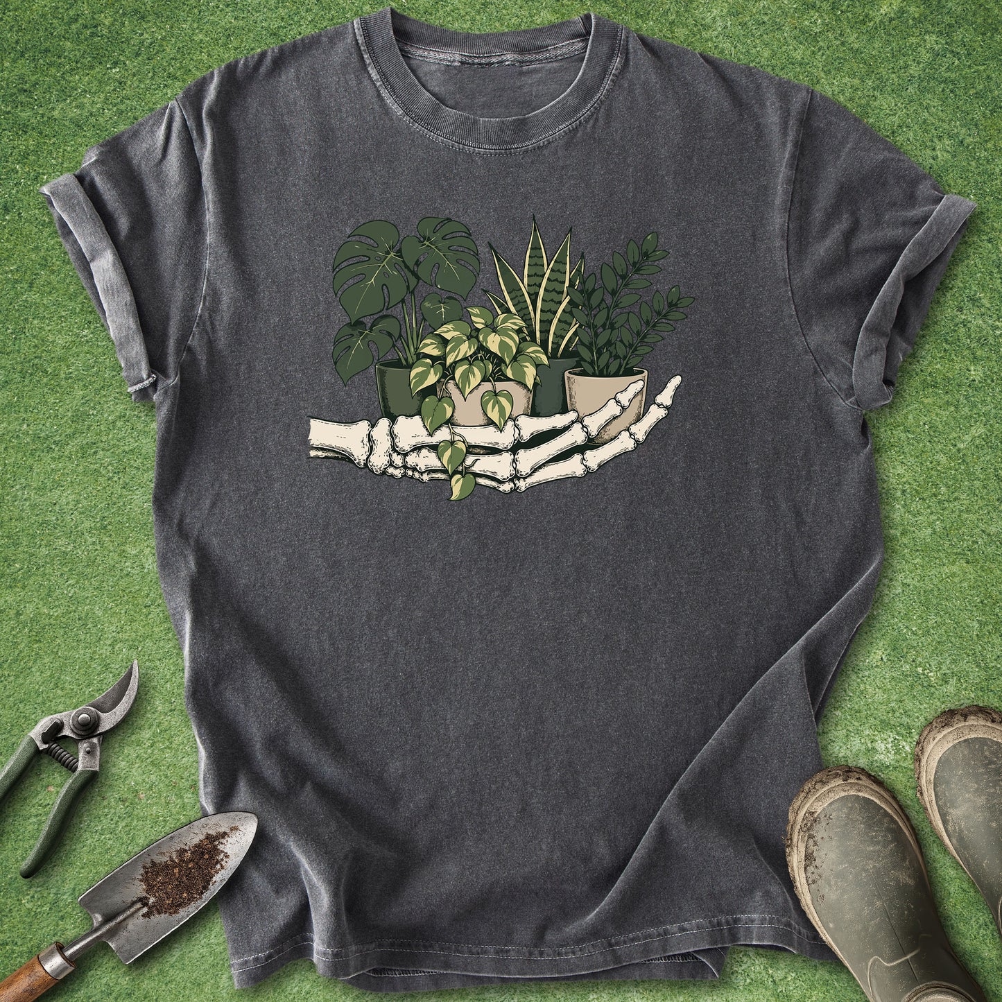 Botanical t-shirt featuring a skeleton hand holding potted Monstera, golden pothos, snake plant, and ZZ plant on a dark charcoal background.