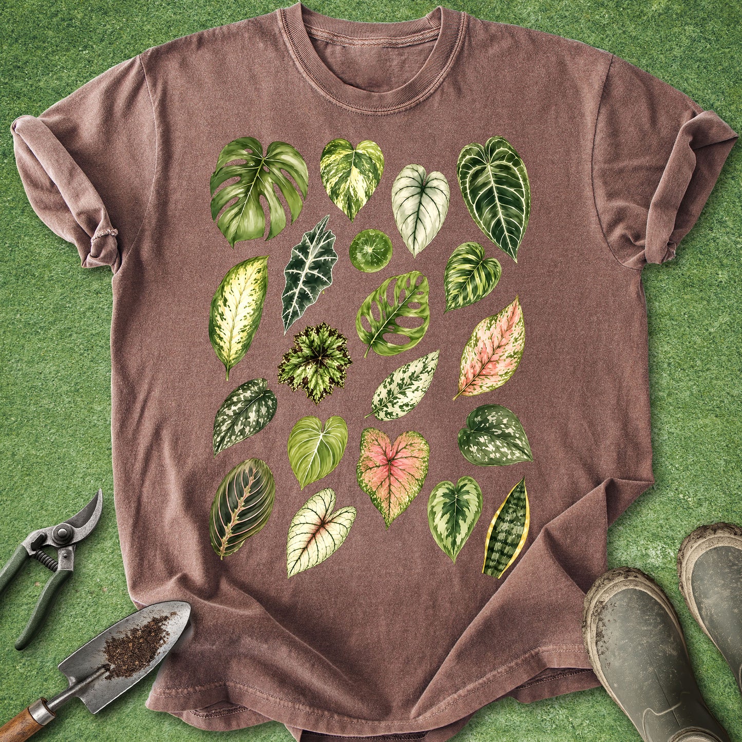 A brown short-sleeve crewneck t-shirt featuring a colorful graphic print of various tropical houseplant leaves, laid flat on a green grass-like surface with gardening tools and boots nearby.