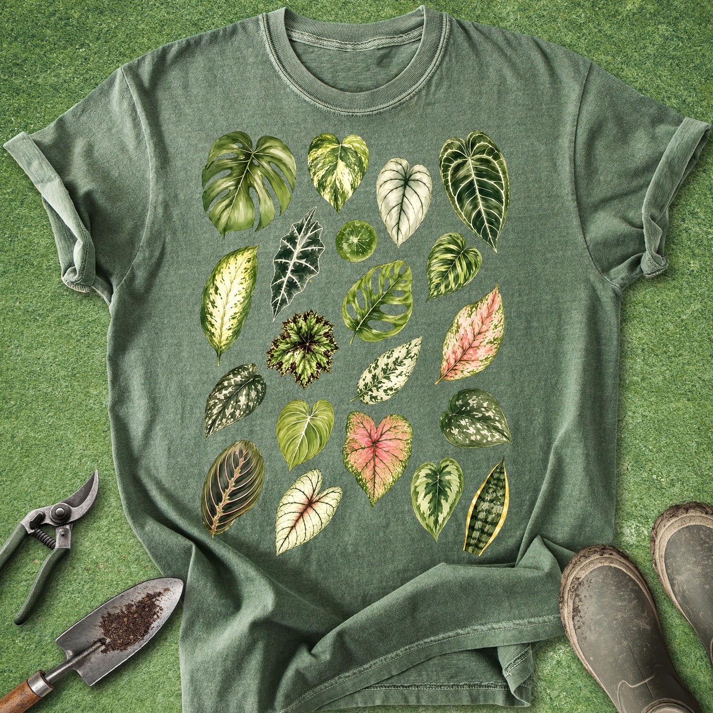 A green short-sleeve t-shirt with a graphic print of various illustrated tropical leaves, styled flat-lay on artificial grass with gardening tools and boots.
