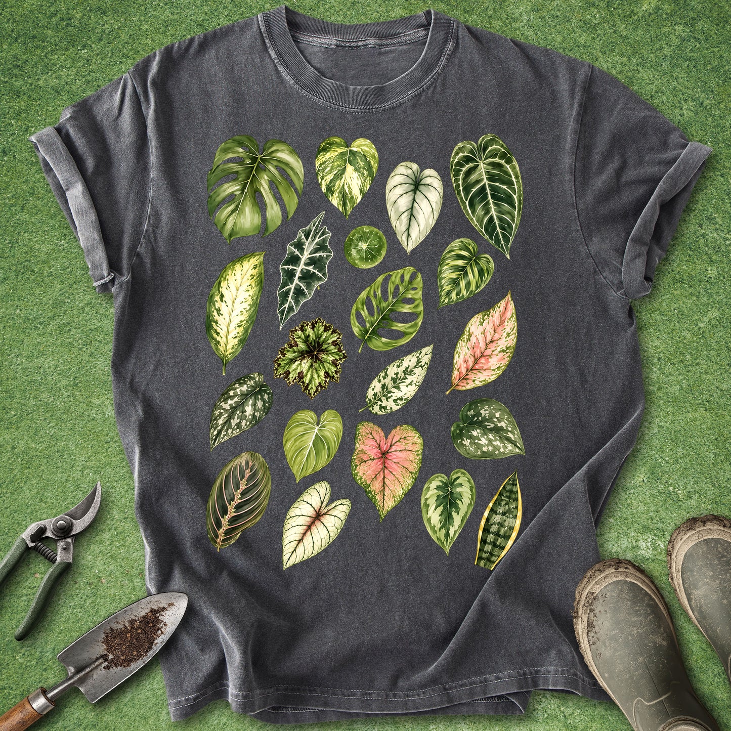 A dark charcoal gray t-shirt with a colorful graphic print of various green houseplant leaves, styled in a flat-lay on artificial green grass with gardening tools and boots.