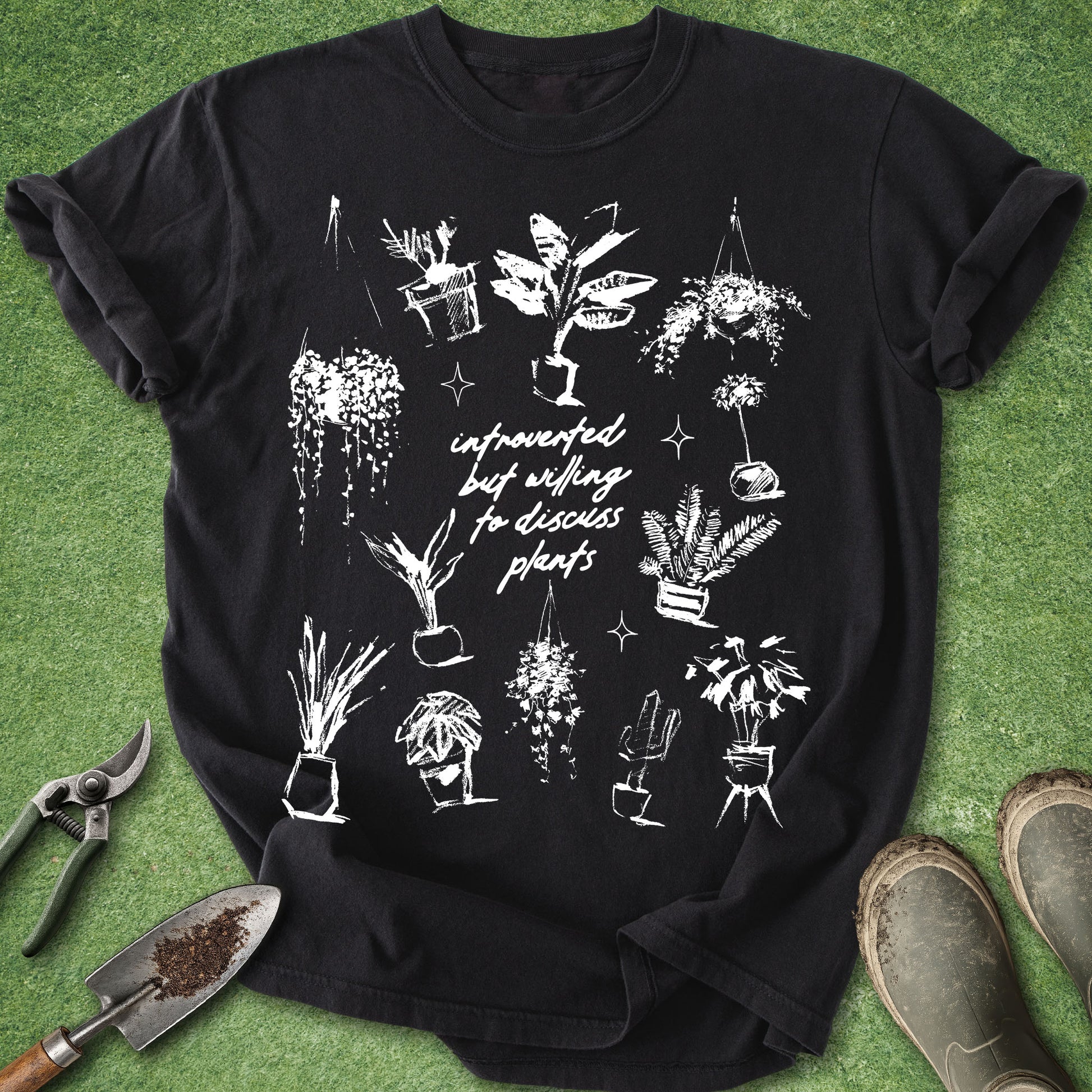 A black short-sleeve graphic T-shirt laid flat on green artificial grass, surrounded by gardening props including pruning shears, a trowel, and boots. The shirt features a white line-art print of various potted plants and the phrase 'infused with will todiscuss plants' in script.