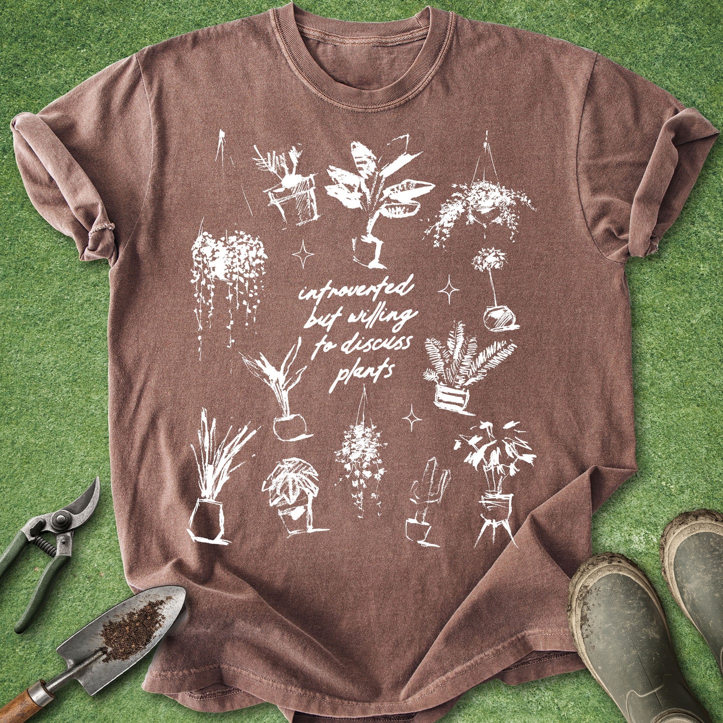 A brown short-sleeve t-shirt with a white hand-drawn style graphic of various potted plants and the text 'infrequently but willinglydiscussing plants' laid flat on green grass with gardening tools and boots nearby.