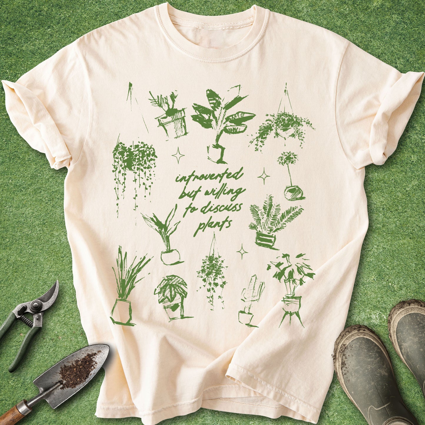 A cream-colored short-sleeve t-shirt with a green graphic print of various houseplants and the text 'unplugged but willing to discuss plants', laid flat on a green grass-like surface with gardening props including pruning shears, a trowel, and boots.