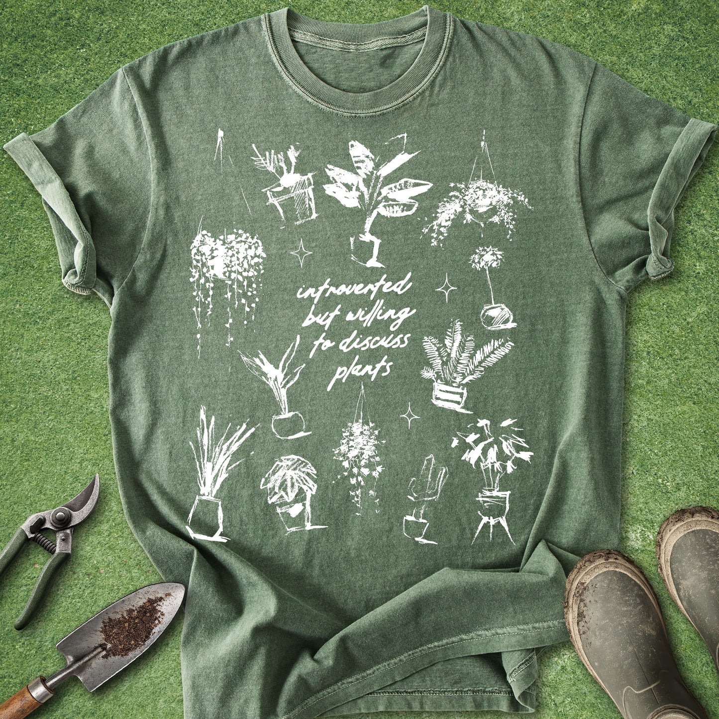 An olive green graphic t-shirt laid flat on artificial grass, surrounded by gardening props including a trowel, pruning shears, and boots. The t-shirt features a white hand-drawn style graphic of various potted plants and the text 'infrequently but willinglydiscussing plants'.